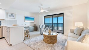 A modern living room with a large window, a dining area, and a kitchen. - OCEAN'S REACH 2X4: 1-Bdrm, Top Floor/Gulf Front Unit! All Amenities Open! (Sanibel Island)