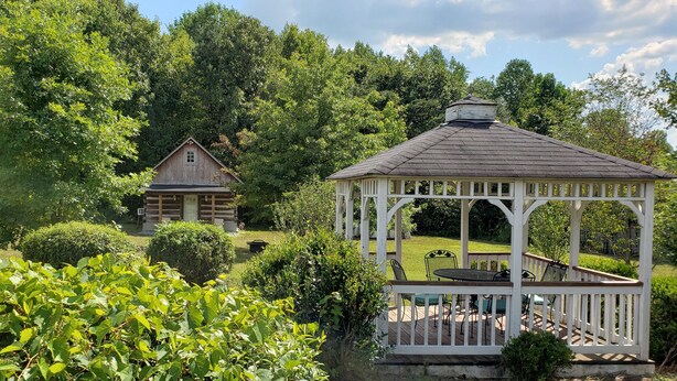 Ein Gazebo mit Holzterrasse und kleinem Tisch, umgeben von üppiger Vegetation und einer Holzhütte in einem Waldgebiet.