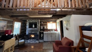 Ein gemütliches Blockhaus-Interieur mit Kamin aus Stein, Holzbalken, einem Schlafzimmer im Zwischengeschoss und einer Küchenzeile.