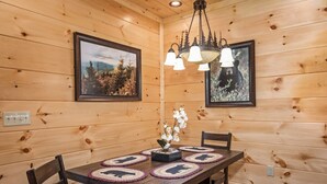 A wooden cabin interior with a dining table, chairs, a chandelier, and framed pictures on the walls.