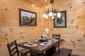 Interior de uma cabana de madeira com uma mesa de jantar, cadeiras, um candelabro e quadros nas paredes.