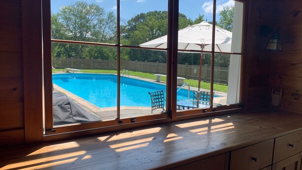 A room with a large window overlooking a swimming pool, wooden cabinets, and a view of a fenced backyard.