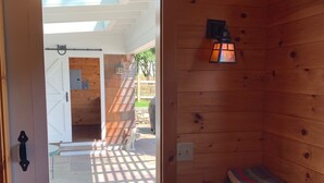 A wooden interior with a door leading to an outdoor area, a small bench with folded towels, and a wall-mounted light fixture.