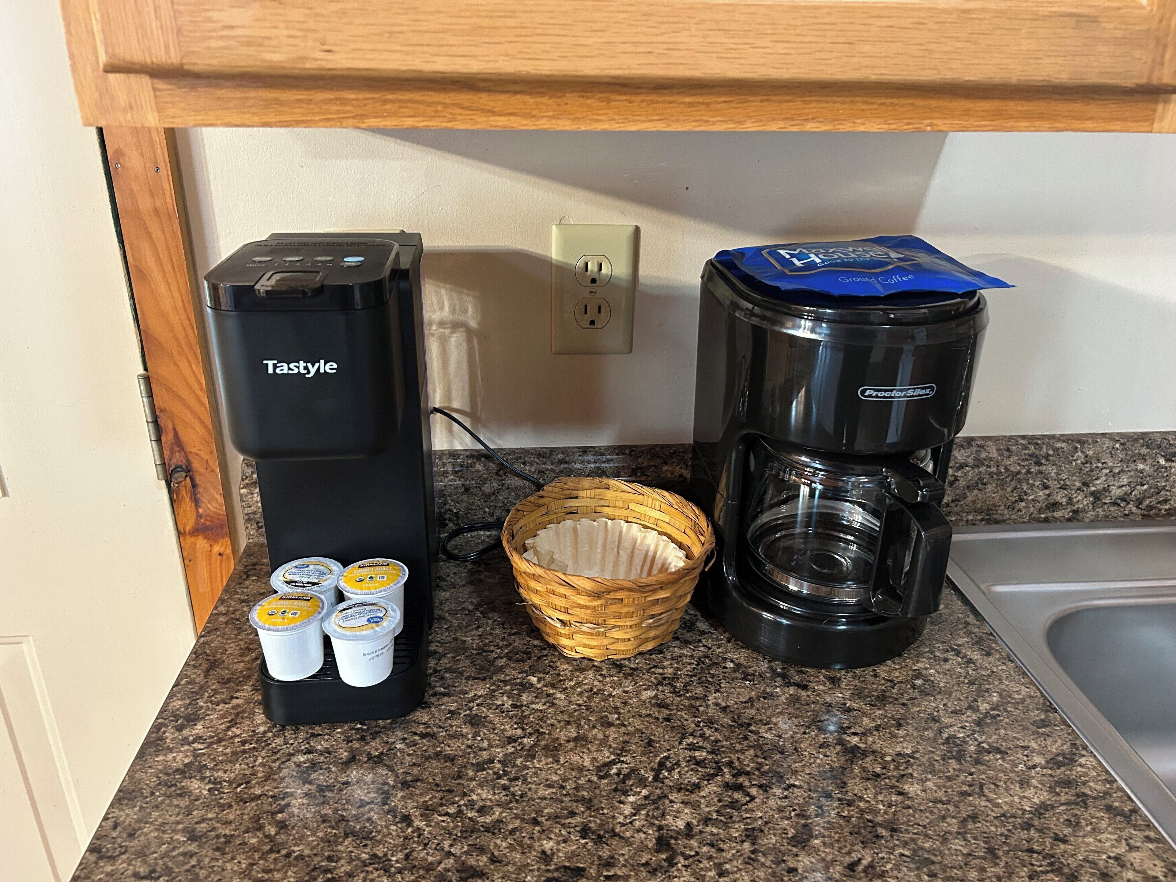 A kitchen with a Tstyle coffee maker, a Proctor-Siler coffee maker, and a basket with coffee pods.