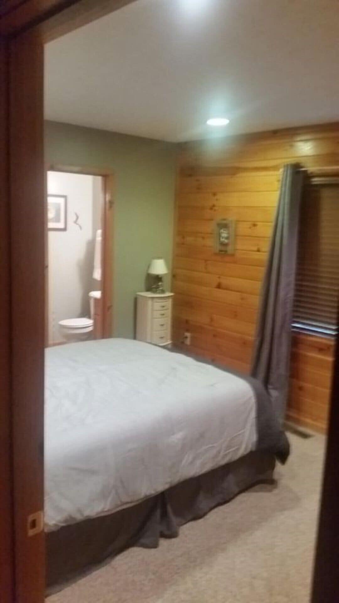 A bedroom with a bed, a small bedside table with a lamp, a wooden wall, and a door leading to a bathroom.