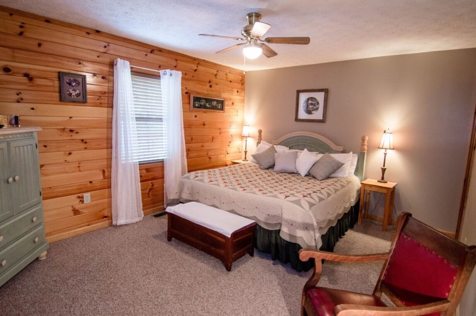 Un dormitorio con pared de madera, una cama con colcha estampada, una mesita de noche con lámpara, un aparador y una silla.