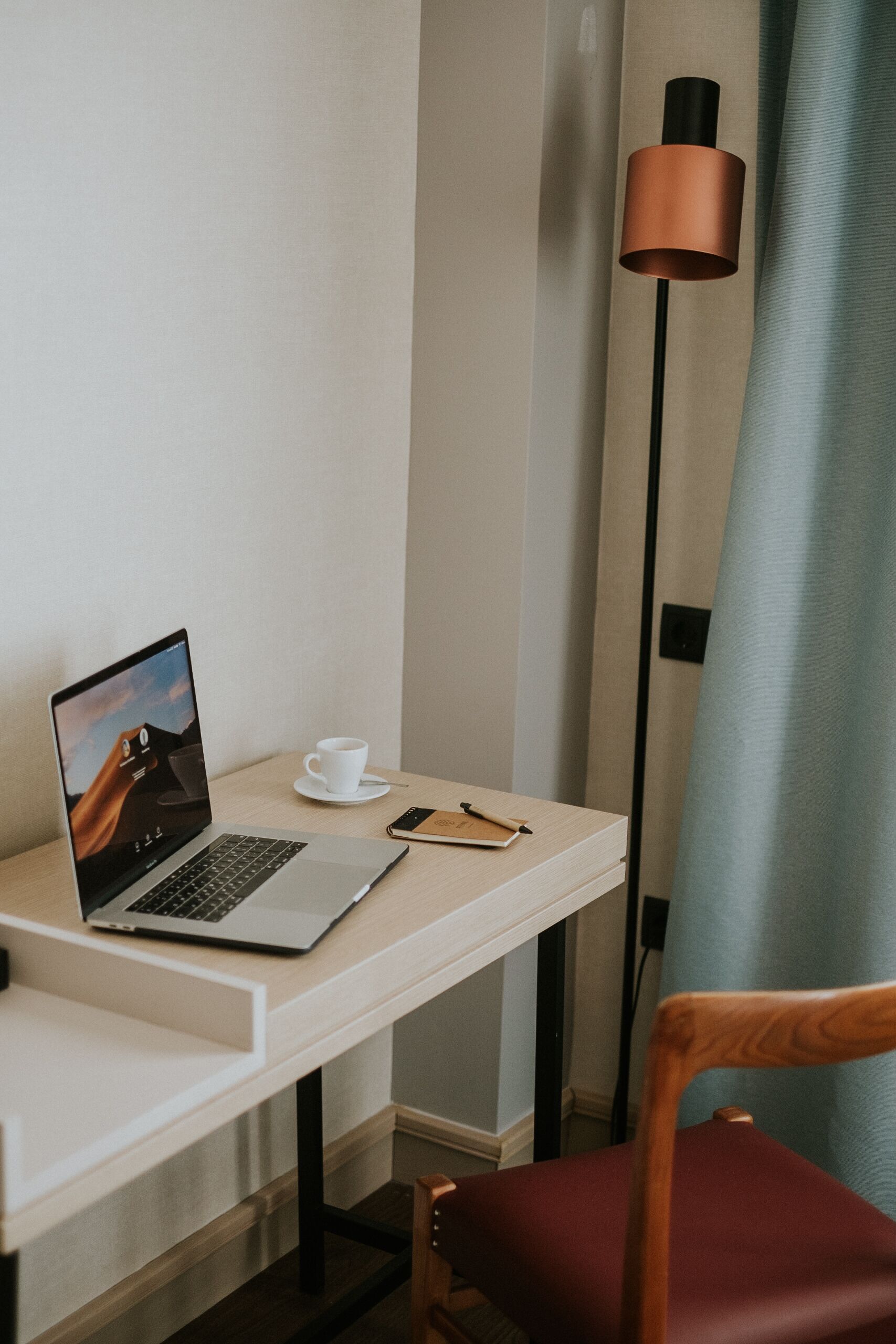 A laptop on a desk with a cup and a notebook, a lamp, and a chair.