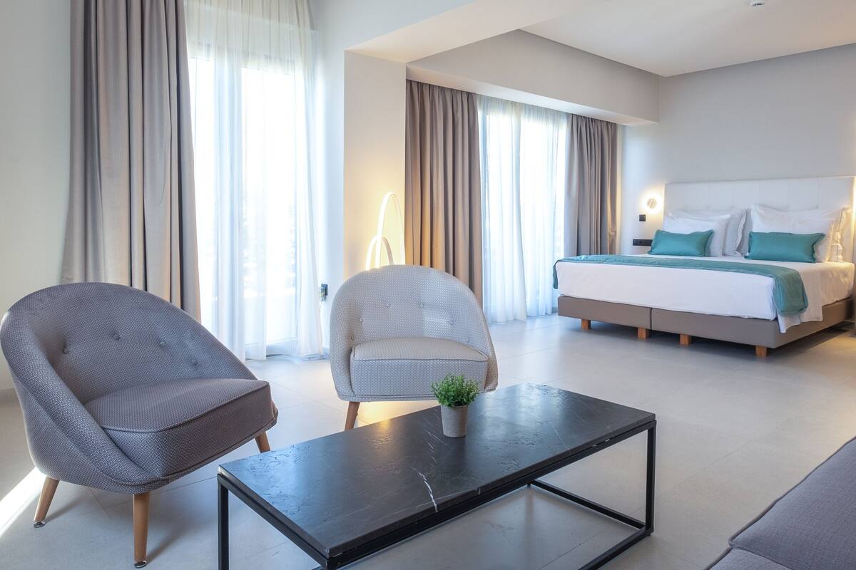 Une chambre d’hôtel moderne dotée d’un grand lit, d’un coin salon avec deux fauteuils, d’une table basse et de grandes fenêtres avec des rideaux.