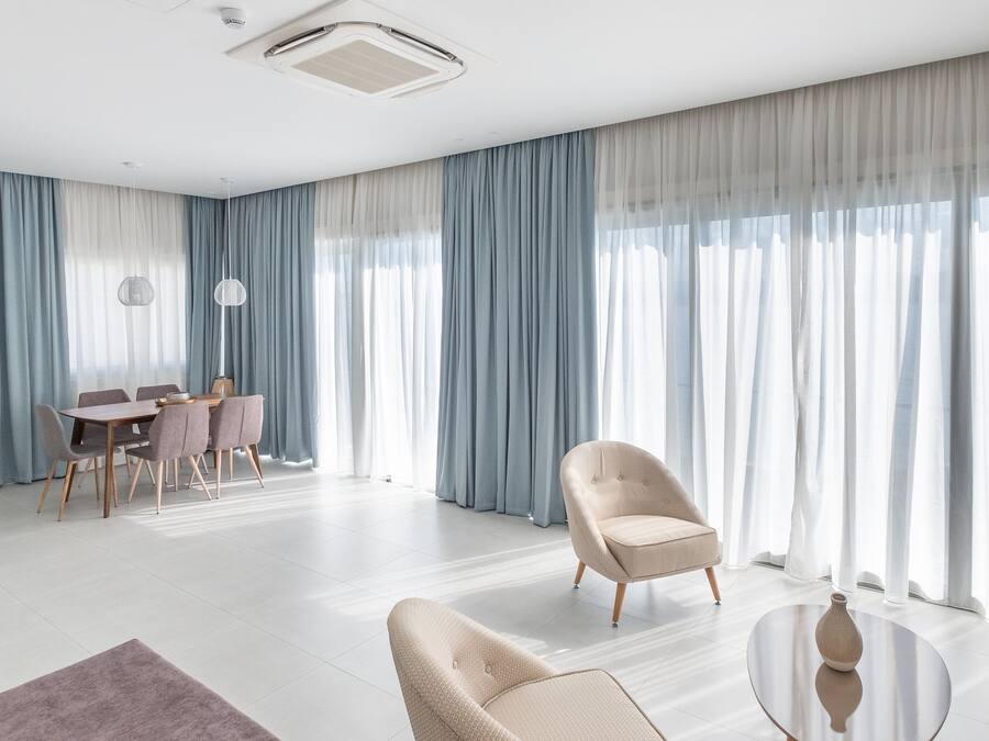 A spacious living room with a modern design, featuring a dining area, two armchairs, a round coffee table, and a large window with sheer curtains.