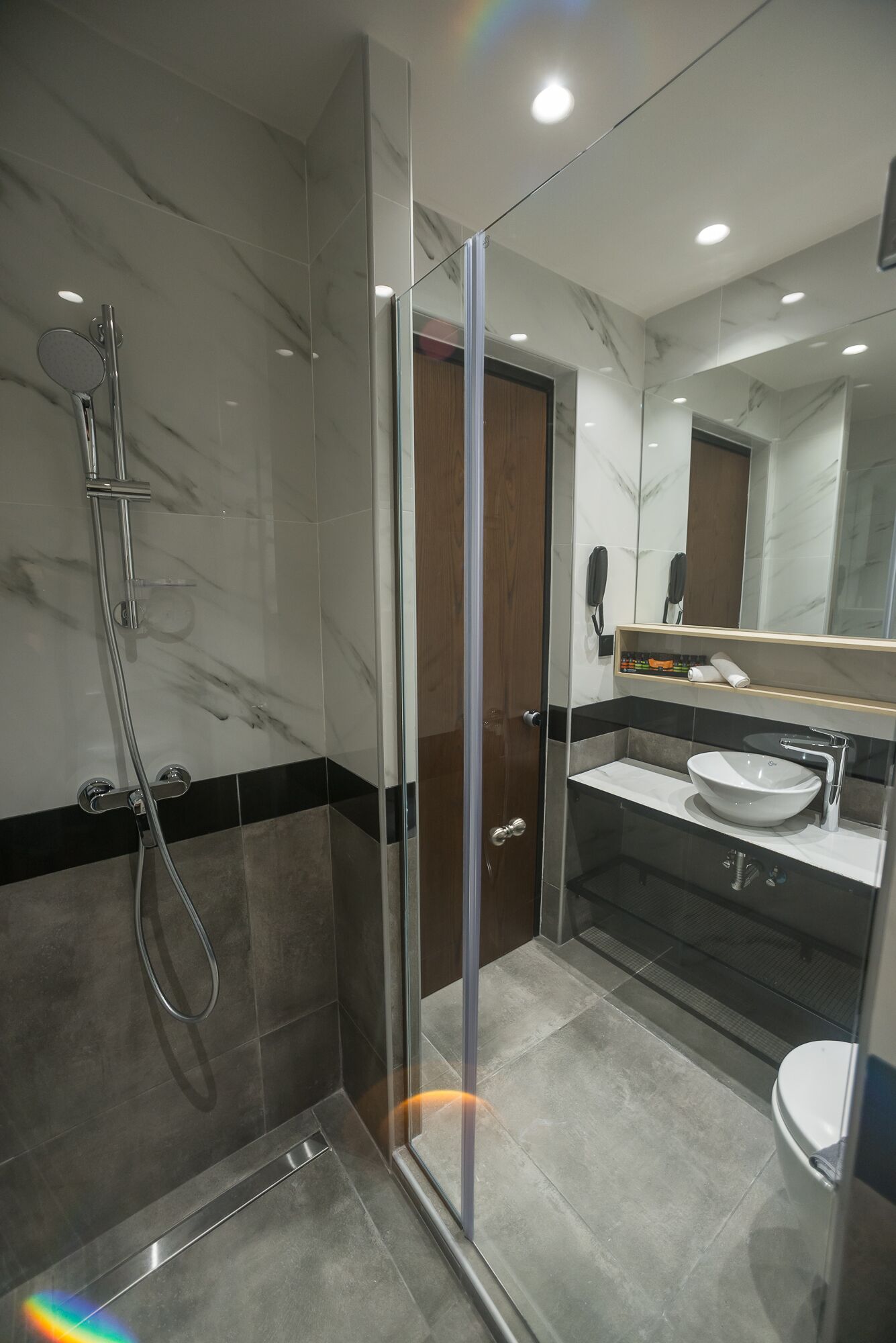 A modern bathroom with a glass-enclosed shower, a marble-tiled wall, a wooden door, and a sink with a mirror above it.