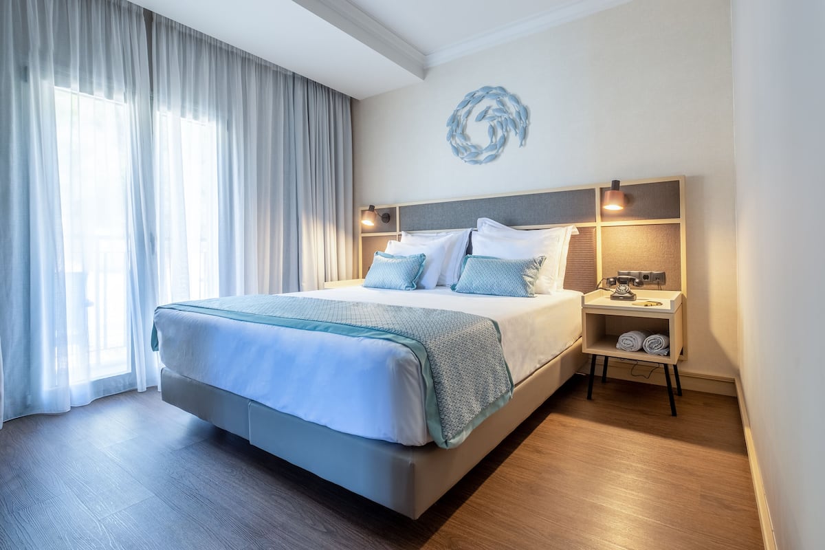 Une chambre d’hôtel moderne avec un grand lit, une table de chevet avec une lampe et une vue sur la ville à travers des rideaux transparents.