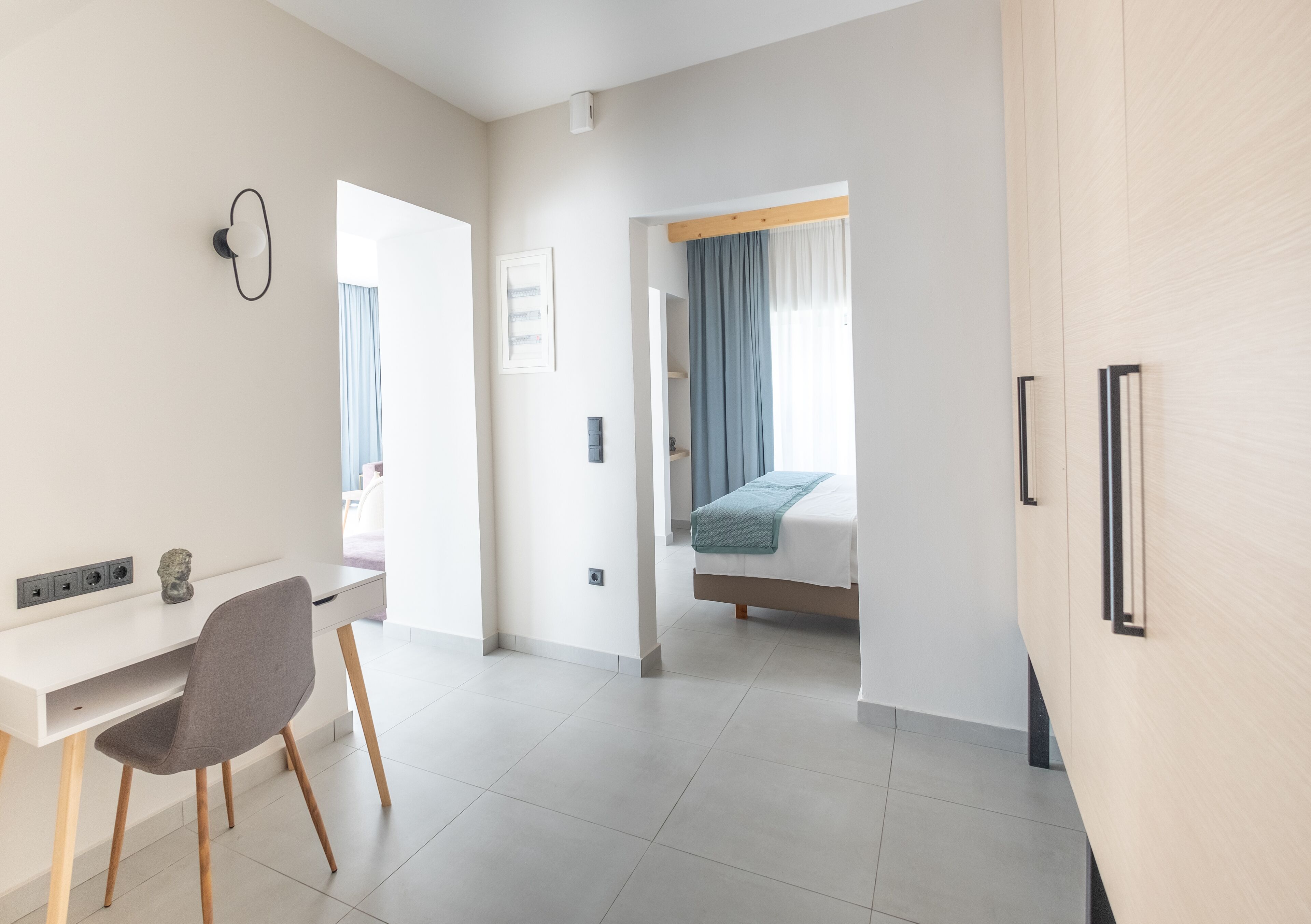 Ett modernt hotellrum med ett vitt skrivbord, en grå stol, en säng med ett blått överkast och en garderob i trä.