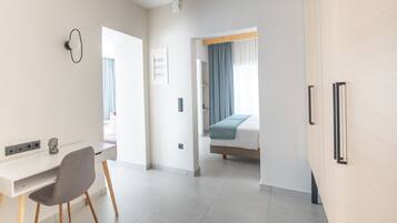 Ett modernt hotellrum med ett vitt skrivbord, en grå stol, en säng med ett blått överkast och en garderob i trä.