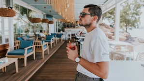 Un hombre sostiene una bebida en un restaurante con mobiliario de madera y grandes ventanales.