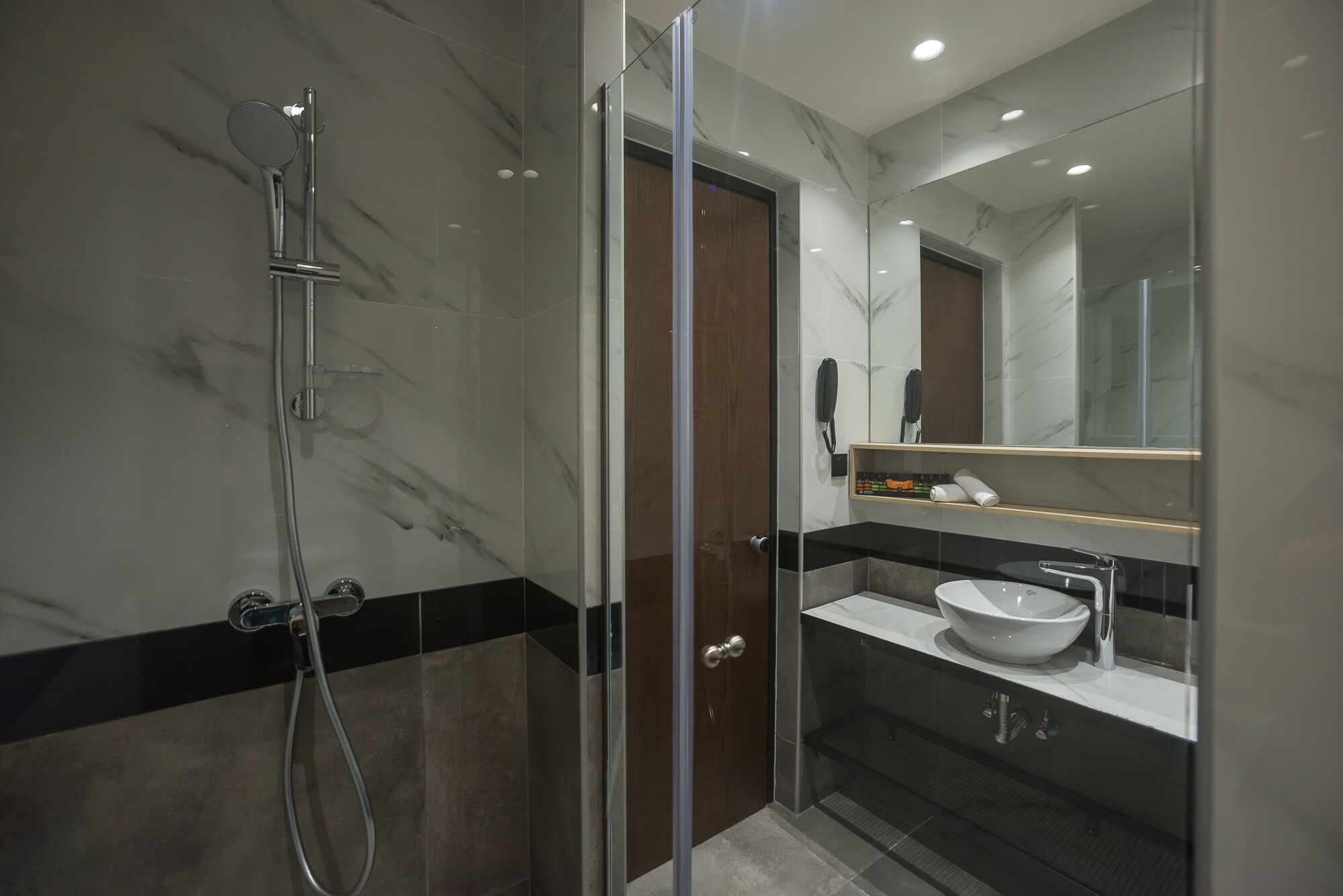 A modern bathroom with a glass-enclosed shower, a wooden door, a marble-tiled wall, a white sink, and a mirror.
