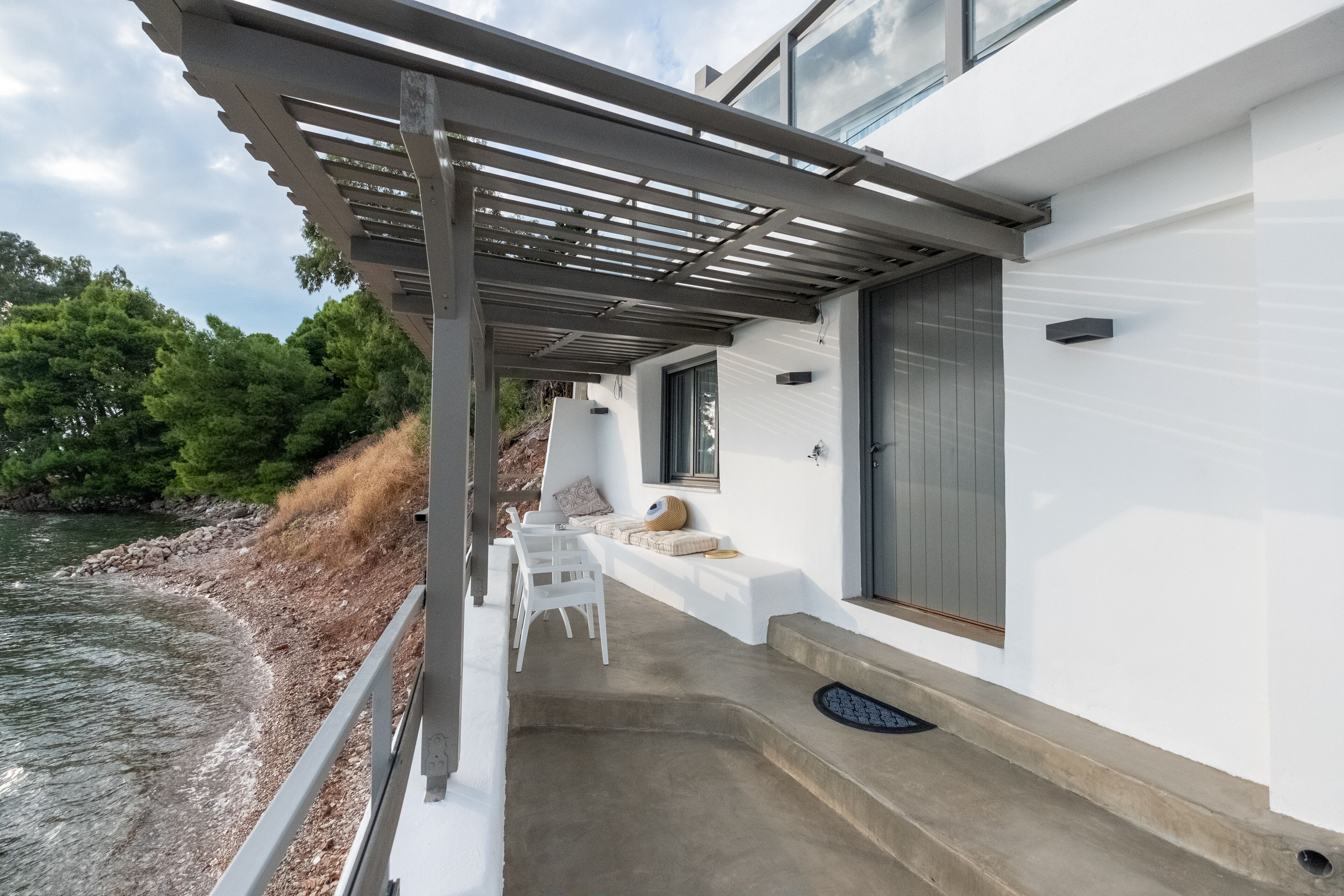 A modern house with a covered patio overlooking a river, featuring a concrete deck, white chairs, and a built-in bench.