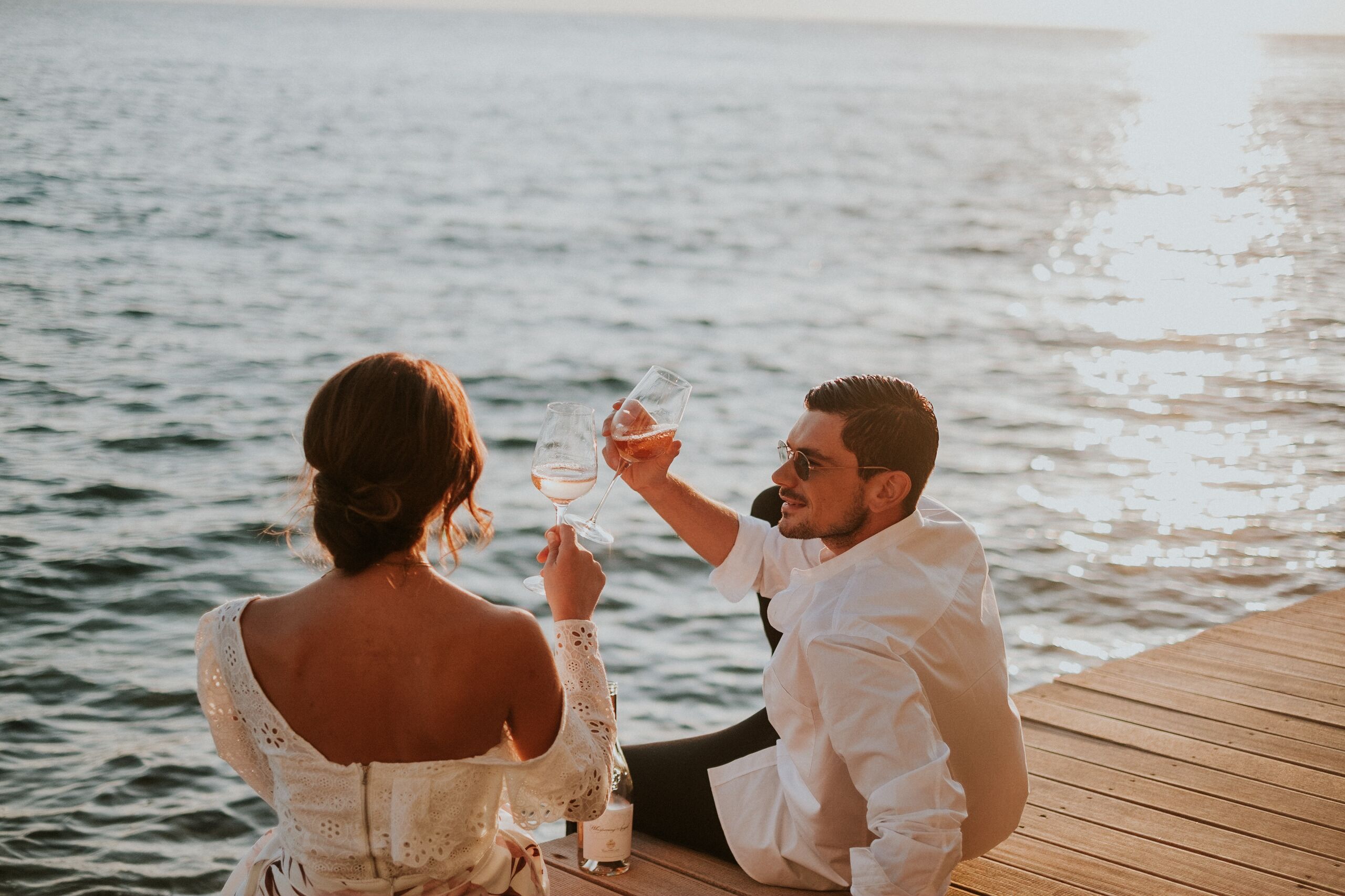 A couple sitting on a wooden dock by the water, holding wine glasses.