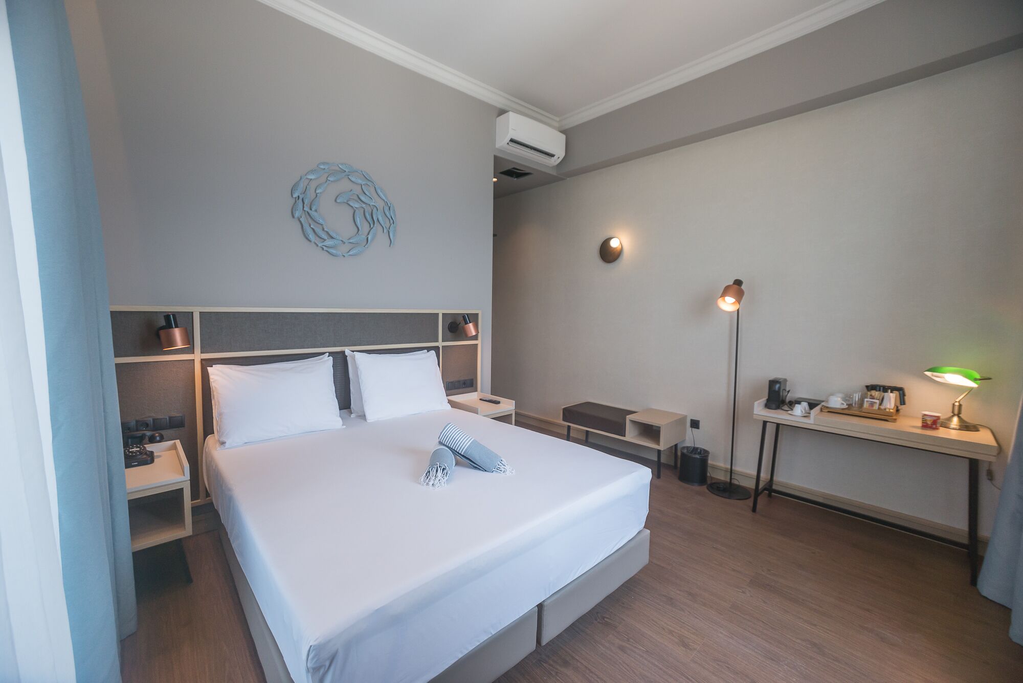A hotel room with a bed, bedside tables, a desk, a lamp, and a wall-mounted air conditioner.