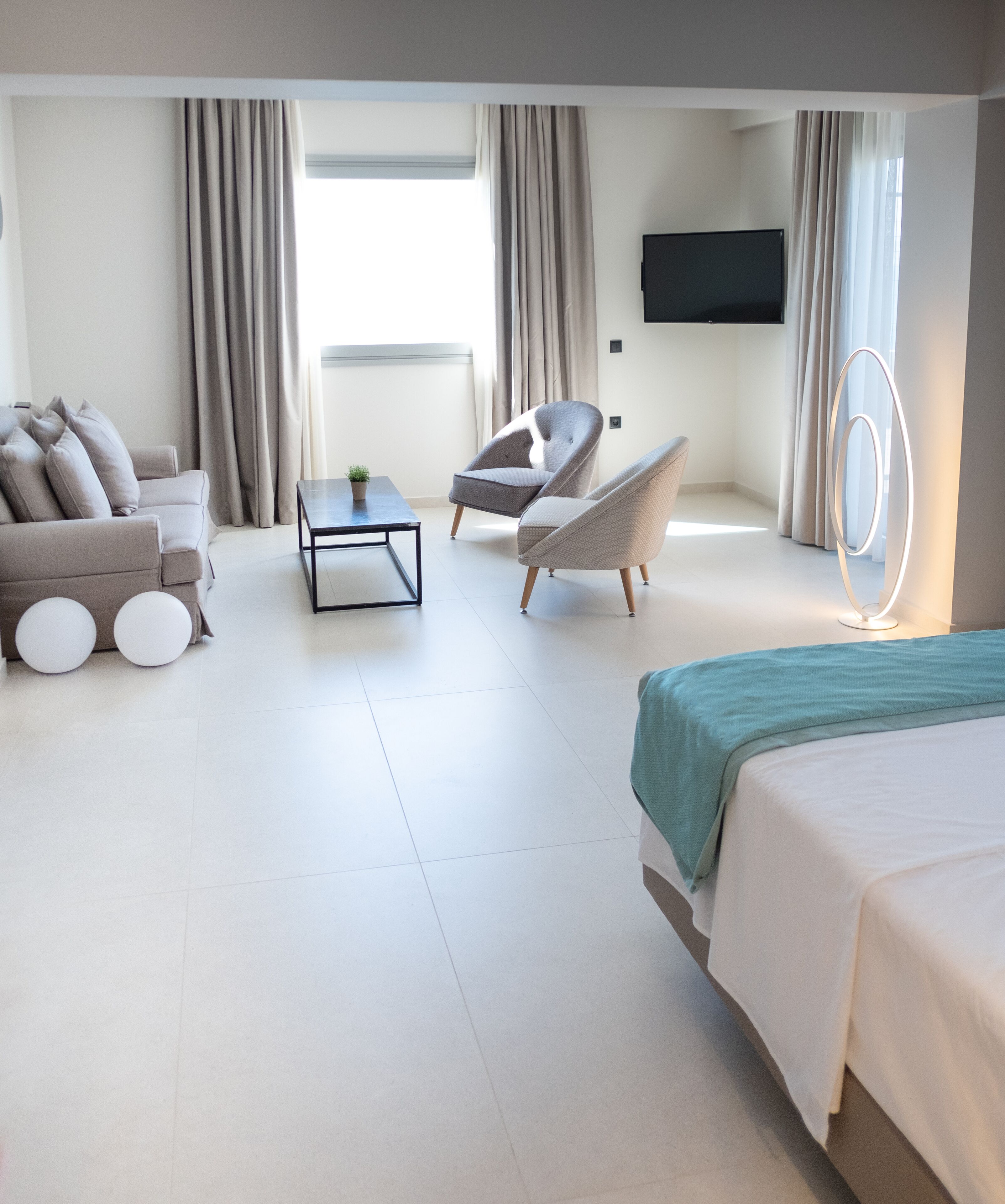 A modern hotel room with a sofa, armchair, and a bed. There is a flat-screen TV mounted on the wall, a small table with a plant, and a unique light fixture.
