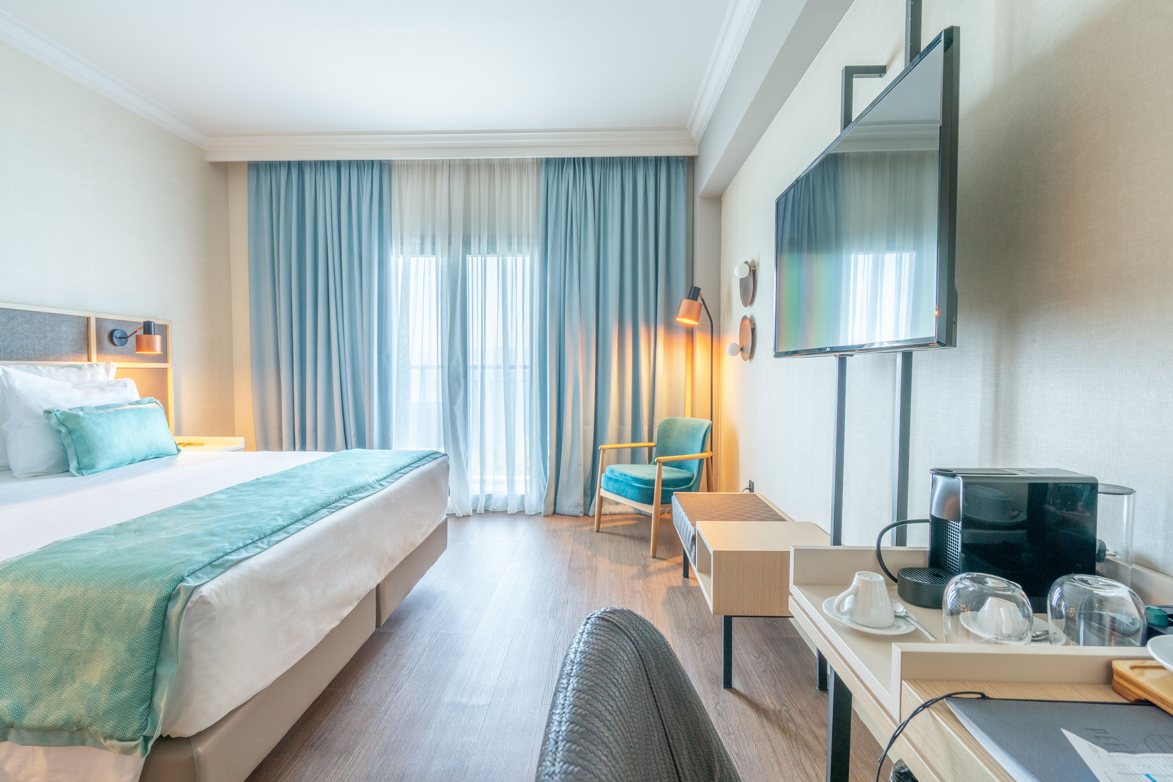 A hotel room with a bed, a desk with a coffee maker, a chair, and a TV on the wall.