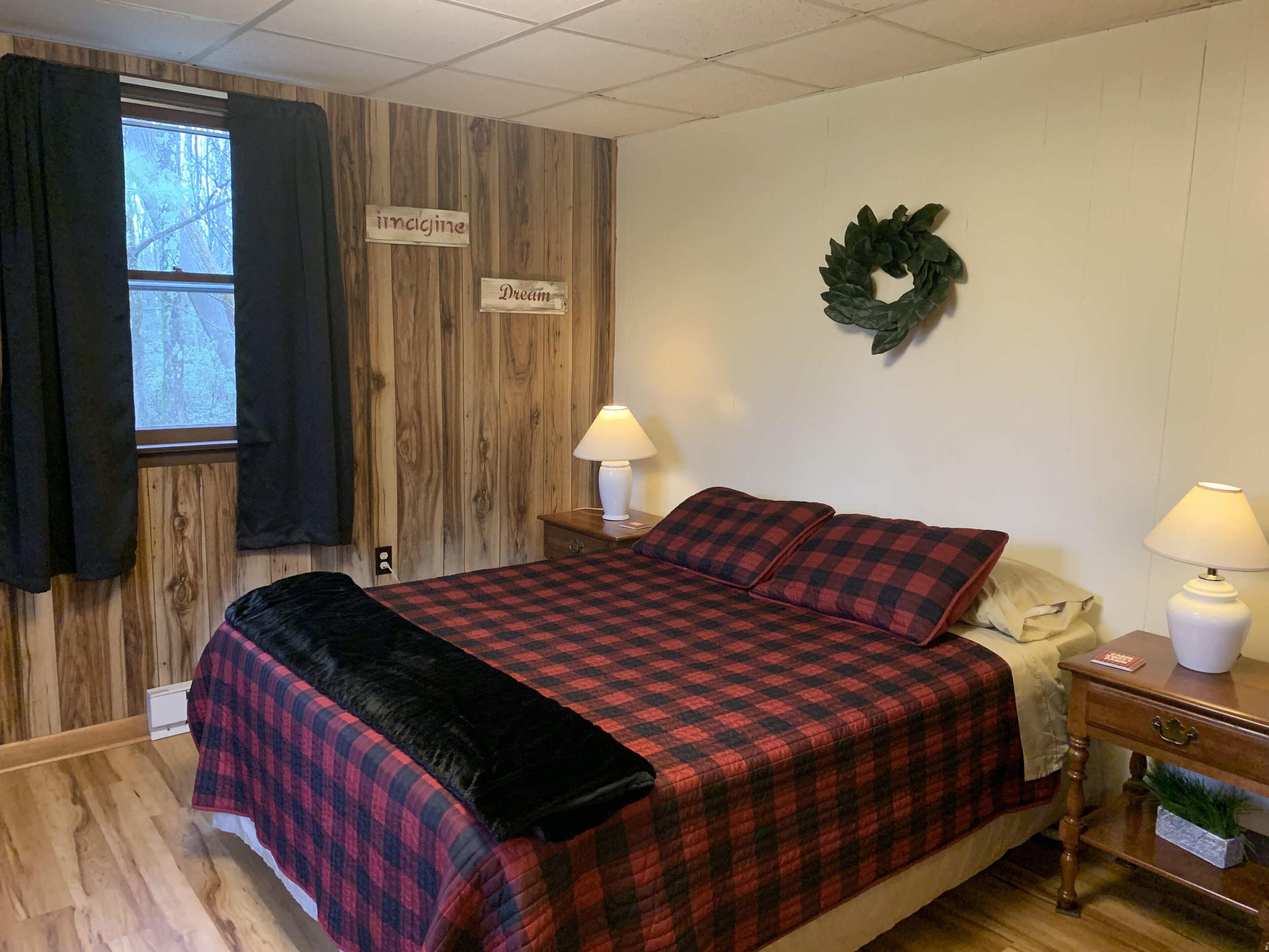 A bedroom with a wooden wall, a bed with a red and black checkered bedspread, two bedside lamps, a nightstand with a lamp, and a wreath on the wall.