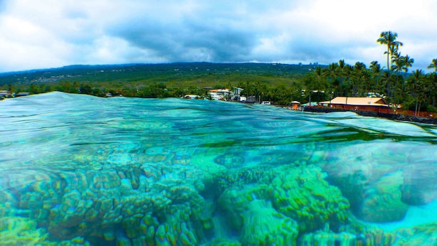 Blick auf die Küste mit klarem türkisblauem Wasser, Korallenformationen und einer fernen Uferlinie mit Gebäuden und Palmen.
