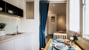 A compact kitchen with a sink, a shelf with pots, a dining area with a table set for two, and a view into the adjacent room. - HAPPYHOMES 102 Luxury Bui Vien Apartment (Ho Chi Minh City)