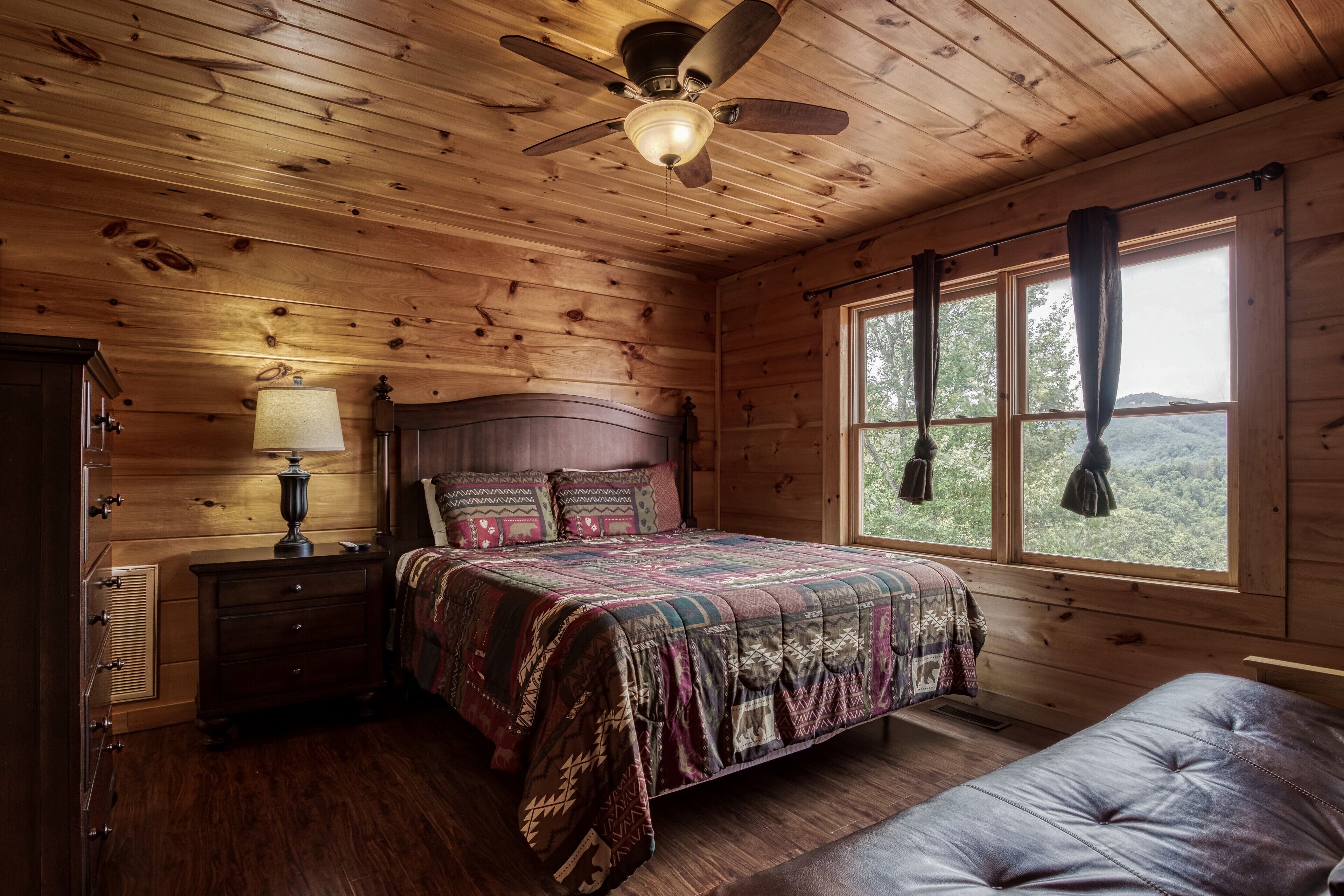 Een slaapkamer in een houten blokhut met een bed, een nachtkastje, een lamp, een plafondventilator en een raam met gordijnen.