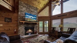 A cozy cabin interior with a stone fireplace, leather sofas, and a large window. - Spacious 4BR Cabin w/ Indoor Pool, Hot Tub, Game Room & Mountain Views (Sevierville)