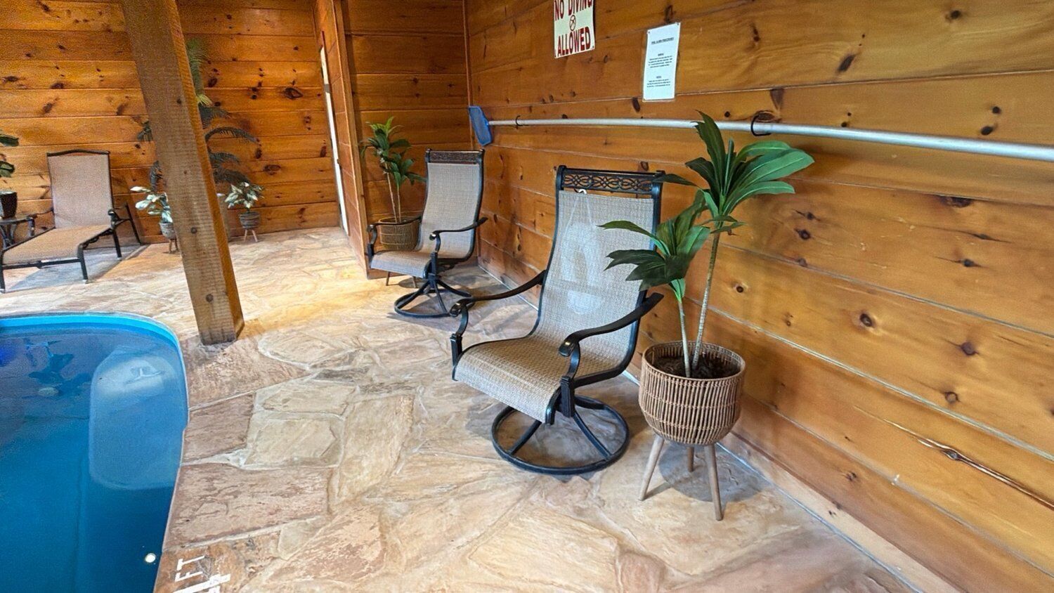 A wooden cabin interior with stone flooring, two cushioned chairs, a small pool, and potted plants.