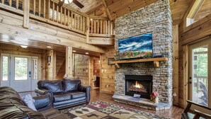 A log cabin interior with a stone fireplace, leather sofas, and a balcony. - Spacious 4BR Cabin w/ Indoor Pool, Hot Tub, Game Room & Mountain Views (Sevierville)