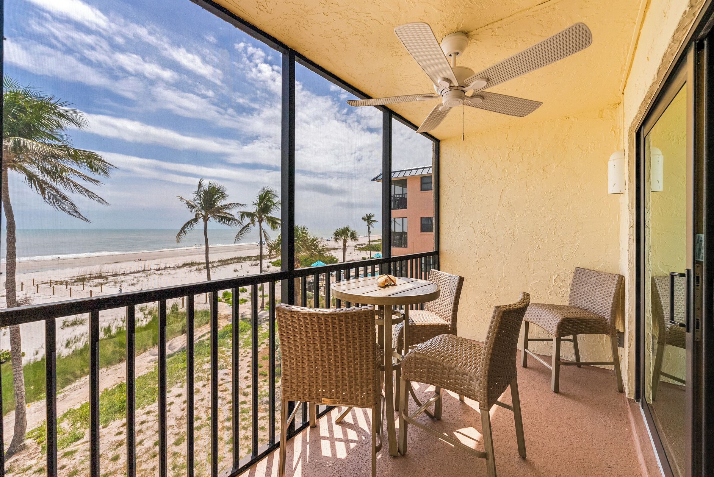 Un balcón con mobiliario de mimbre, ventilador de techo y vista a la playa y palmeras.