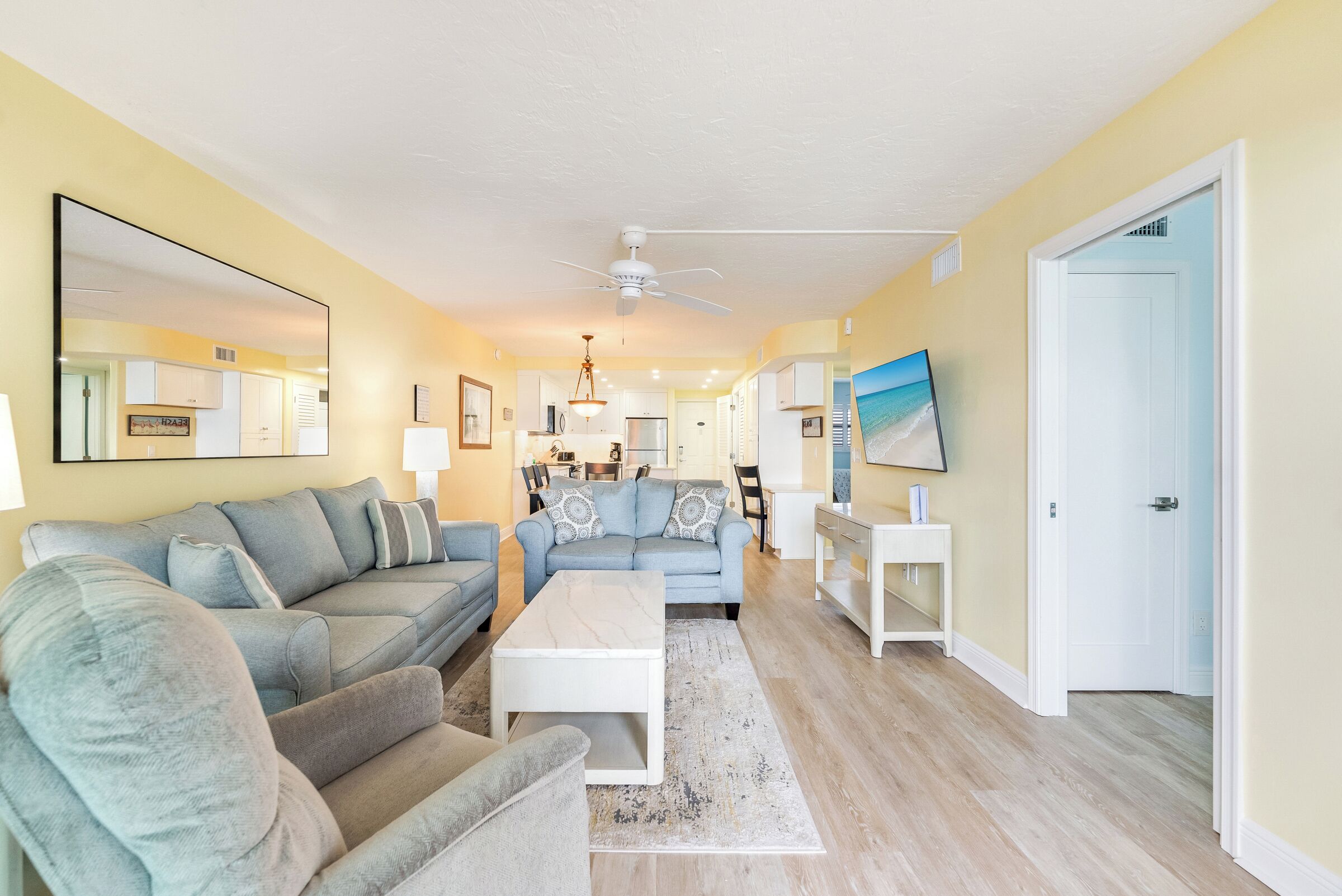 A living room with a sofa, armchair, coffee table, and a TV. There is a kitchen in the background.