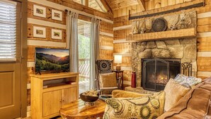 A cozy cabin interior with a stone fireplace, wooden walls, a television, and a comfortable seating area.