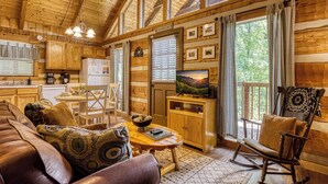 A cabin interior with a wooden ceiling, a dining area, a living room with a sofa and a rocking chair, and a kitchen with wooden cabinets and a refrigerator.