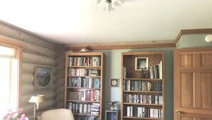 Um recanto acolhedor para leitura, com uma poltrona de couro, uma estante de madeira repleta de livros e uma mesinha com abajur e flores.
