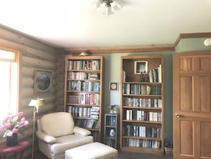 Cozy cabin-style room features a comfortable armchair, bookshelf, and small table with a lamp and vase of flowers.