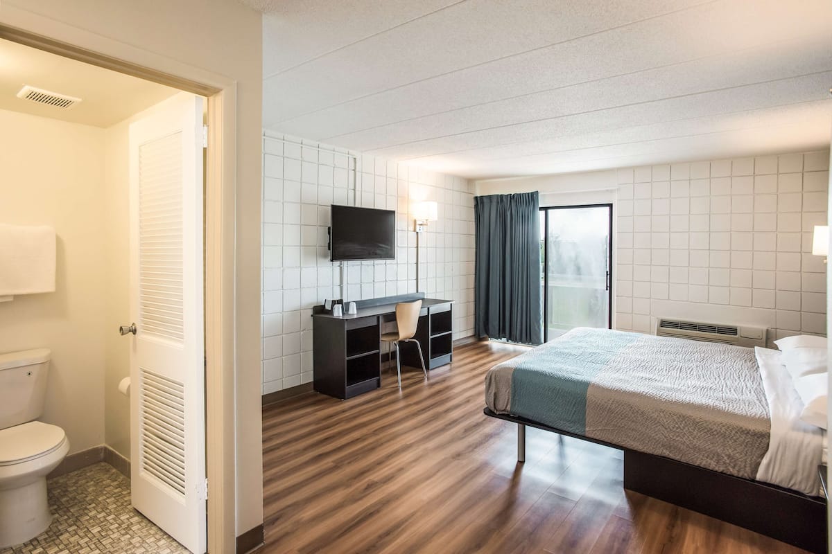 A hotel room with a wooden floor, a bed, a desk with a chair, a television, and a bathroom with a toilet and shower.