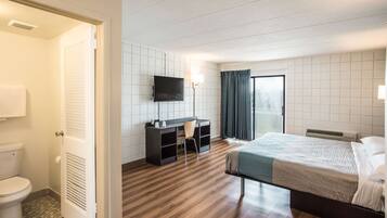 A hotel room with a wooden floor, a bed, a desk with a chair, a television, and a bathroom with a toilet and shower.