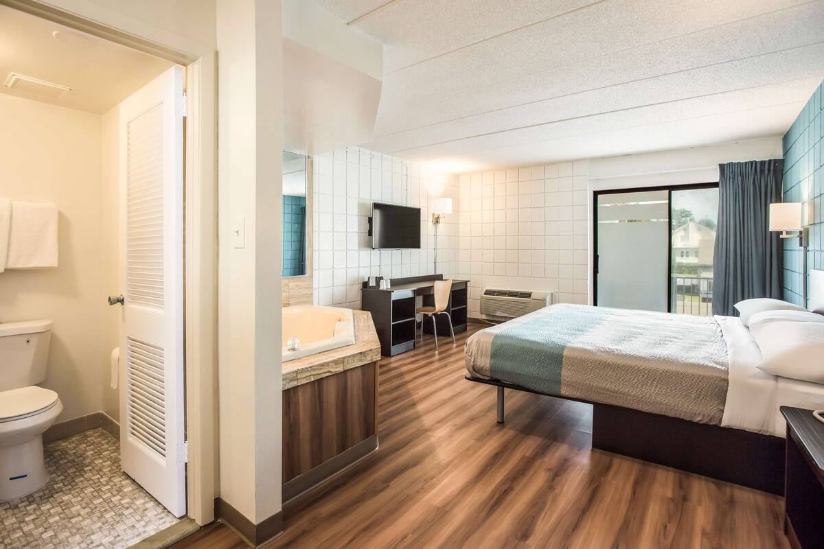 A hotel room with a wooden floor, a bed, a desk, a chair, a TV, a bathroom with a toilet, and a shower.