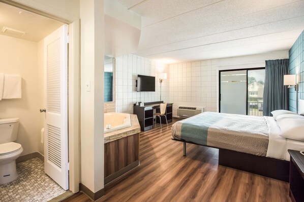 A hotel room with a wooden floor, a bed, a desk, a chair, a TV, a bathroom with a toilet, and a shower.