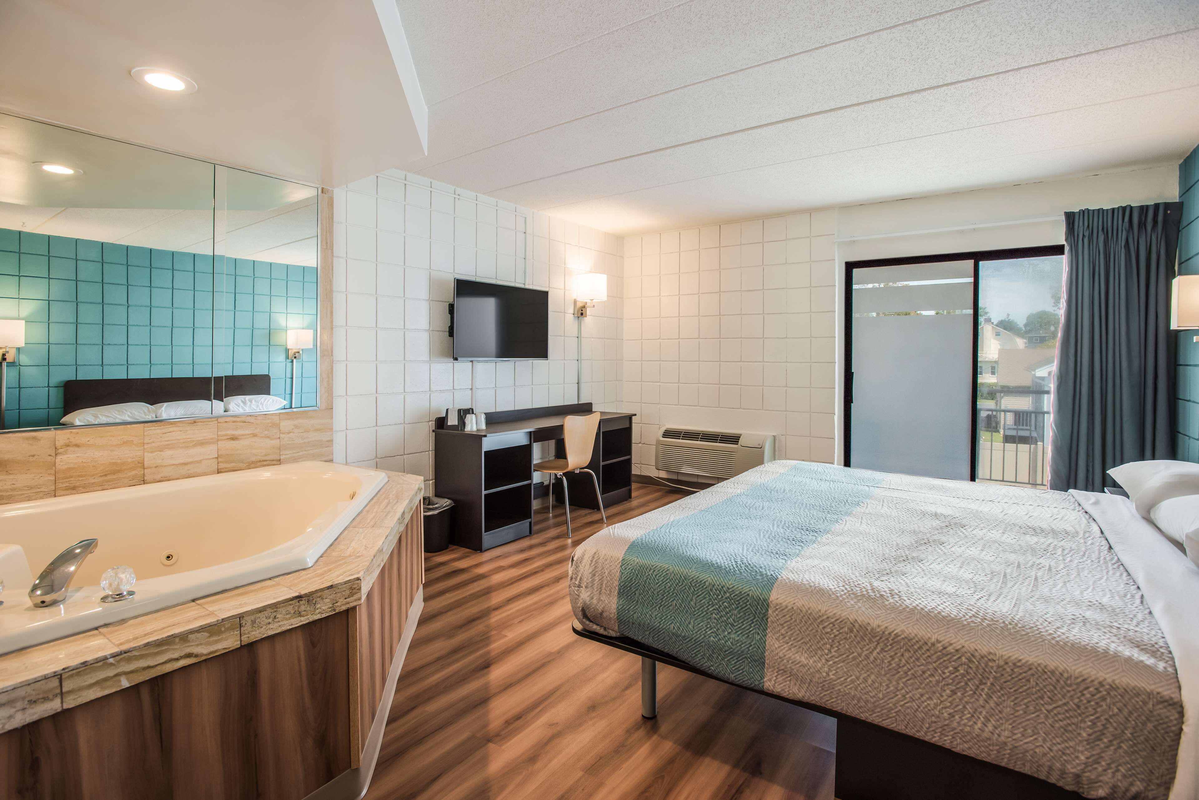 Habitación de hotel con una bañera grande, una cama con colcha, un escritorio con silla, un televisor montado en la pared y una puerta corrediza de vidrio al balcón.