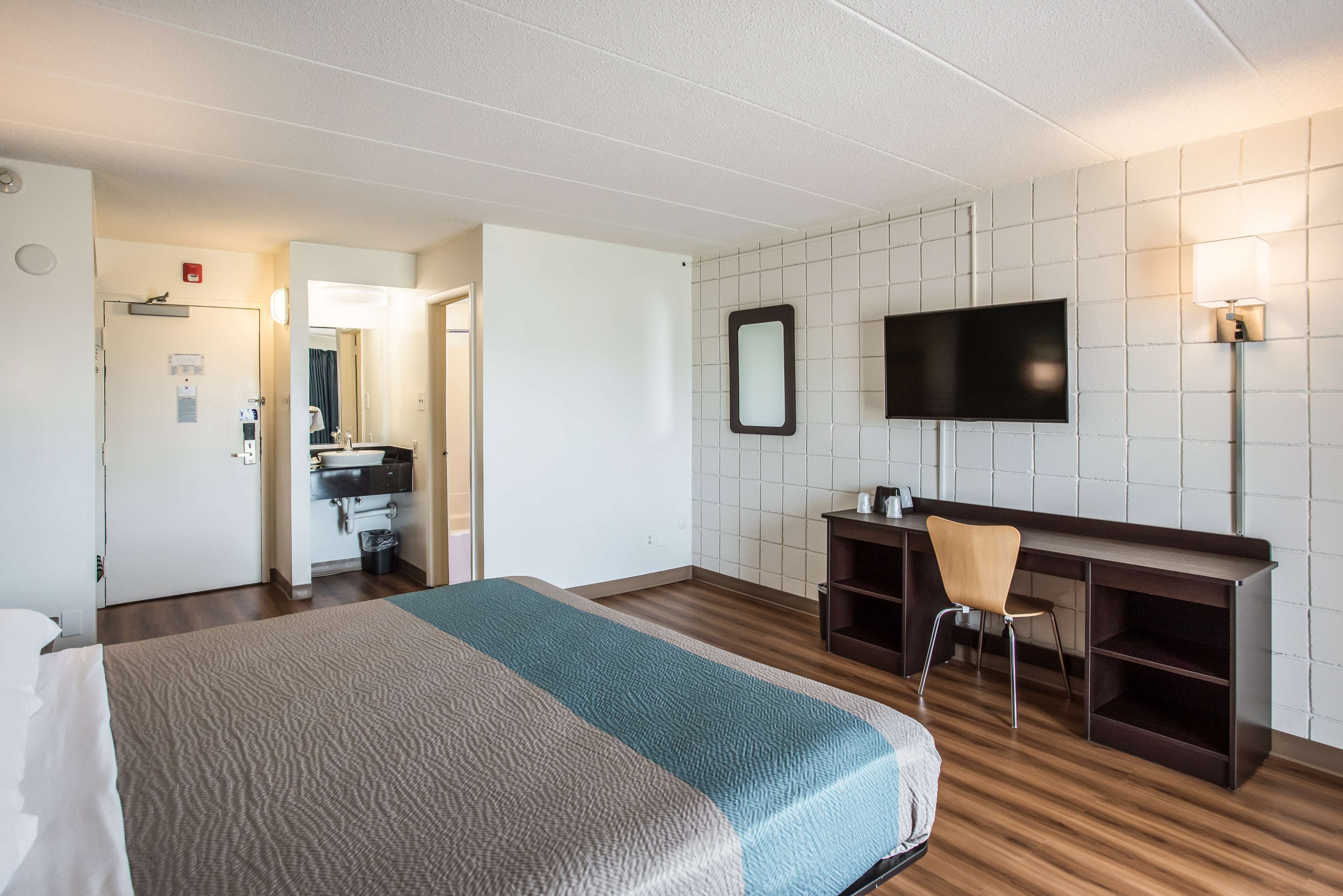 A hotel room with a bed, a desk, a chair, a TV, and a bathroom with a sink and mirror.