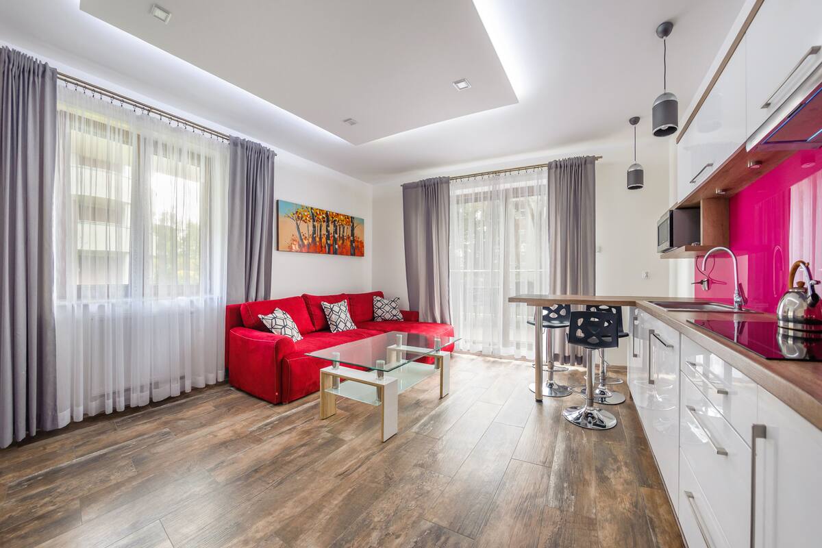 A modern living room with a red sofa, a glass coffee table, and a kitchen area with a pink cabinet and a microwave.