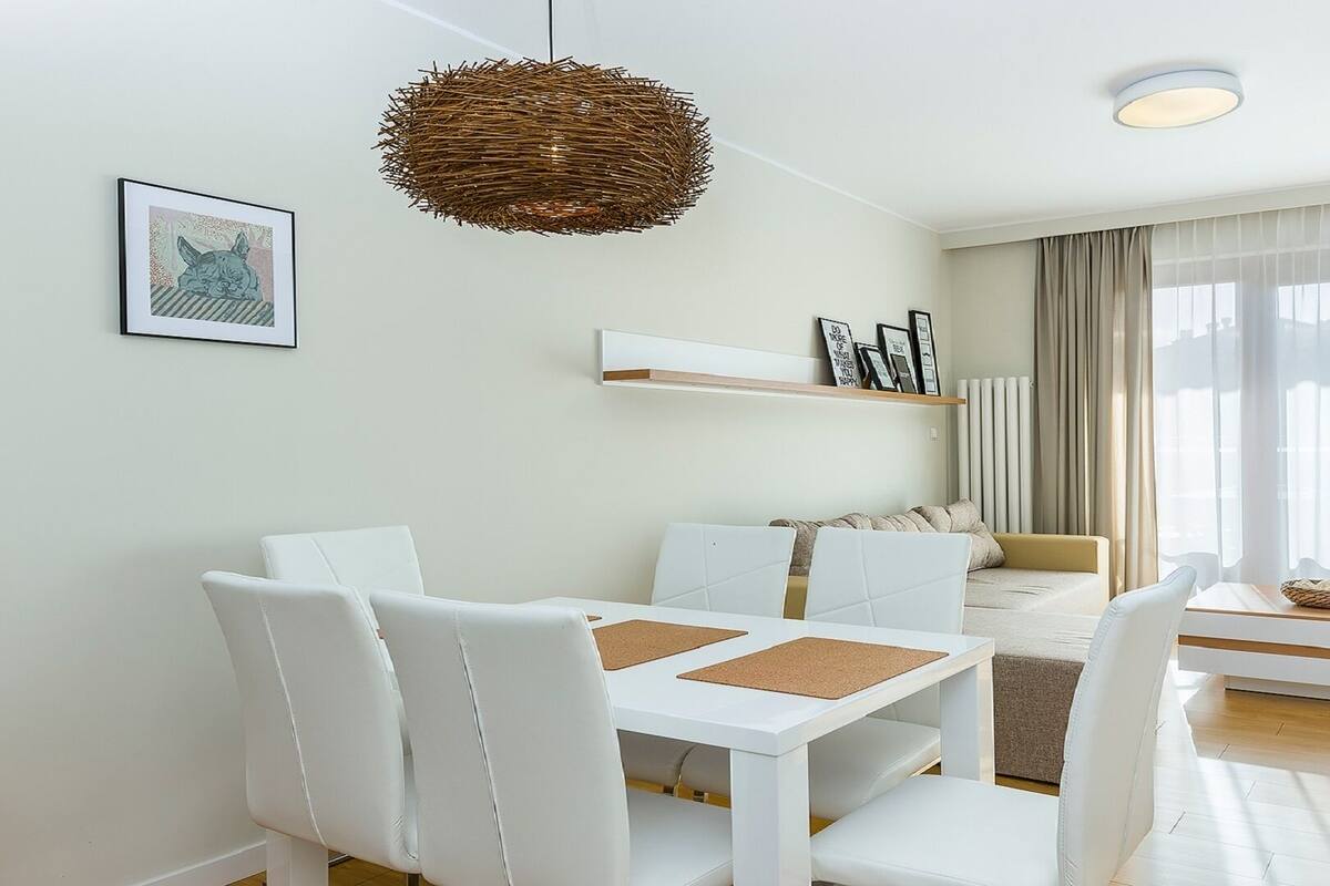A modern living room with a white sofa, a dining table with white chairs, a woven pendant light, and a large window with sheer curtains.