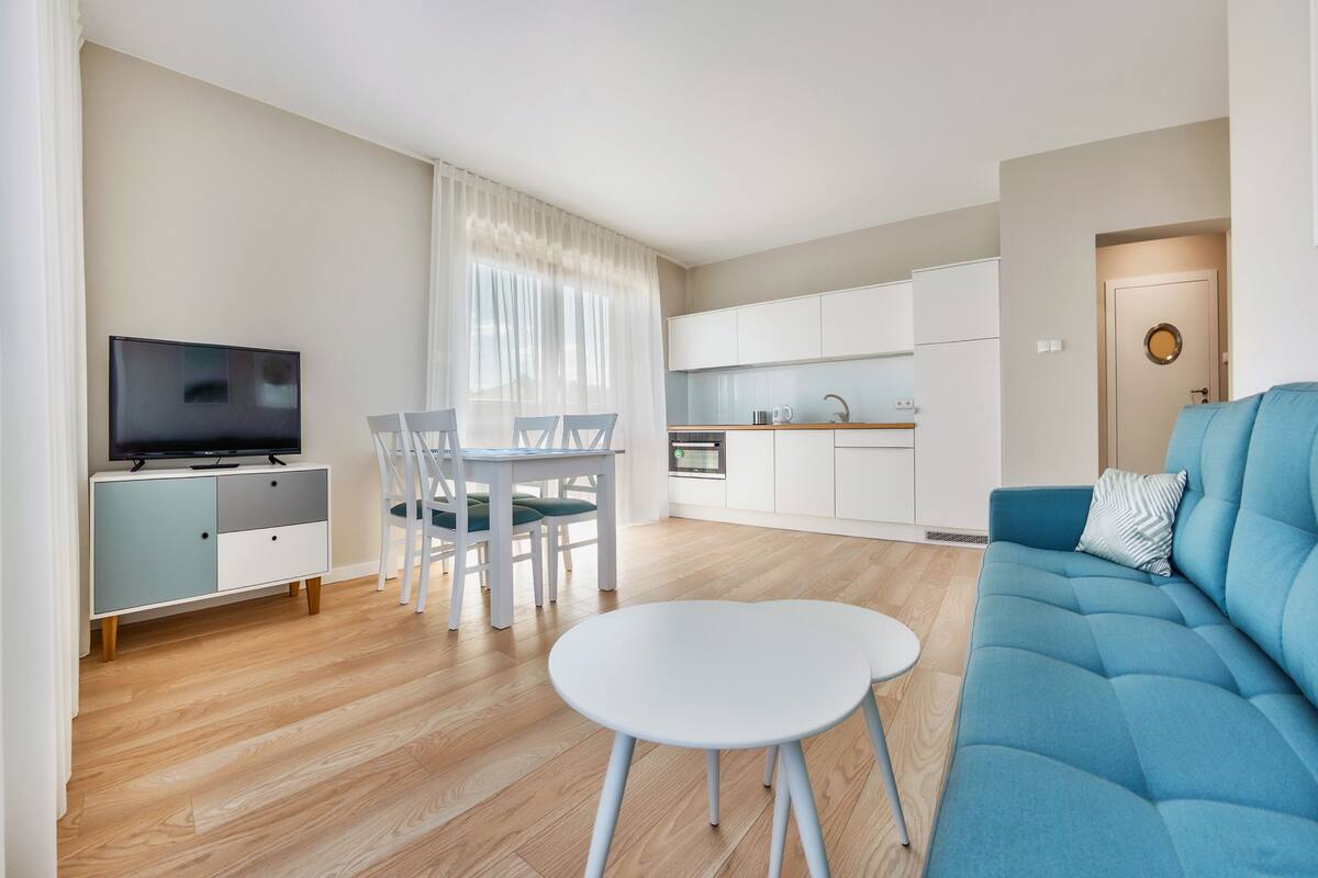 A modern living room with a blue sofa, a white dining table, and a kitchen area with white cabinets and a built-in oven.