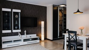Een moderne woonkamer met een flatscreen televisie aan de muur, een witte entertainmentkast met glazen planken, een hoge vloerlamp en een eethoek met een tafel en stoelen.