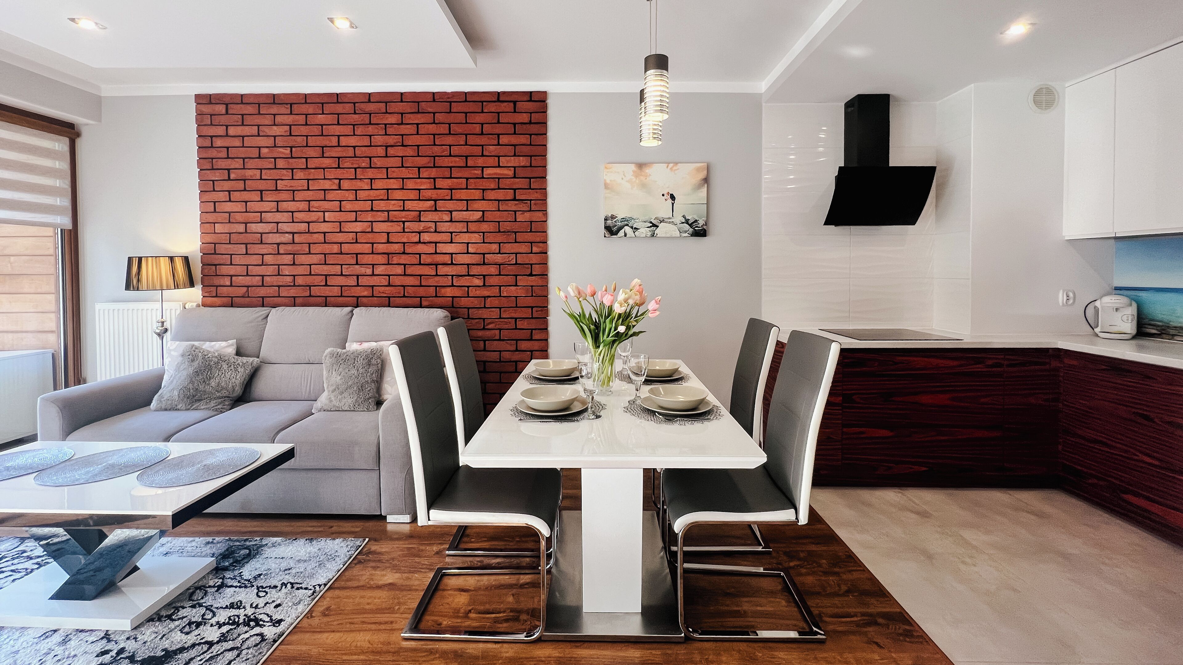 A modern living room with a grey sofa, a dining table set with plates and glasses, and a brick accent wall.