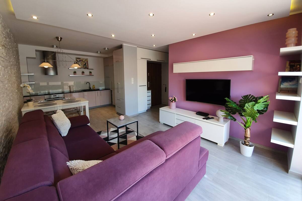 A modern living room with a purple sofa, a TV mounted on the wall, and a kitchen in the background.