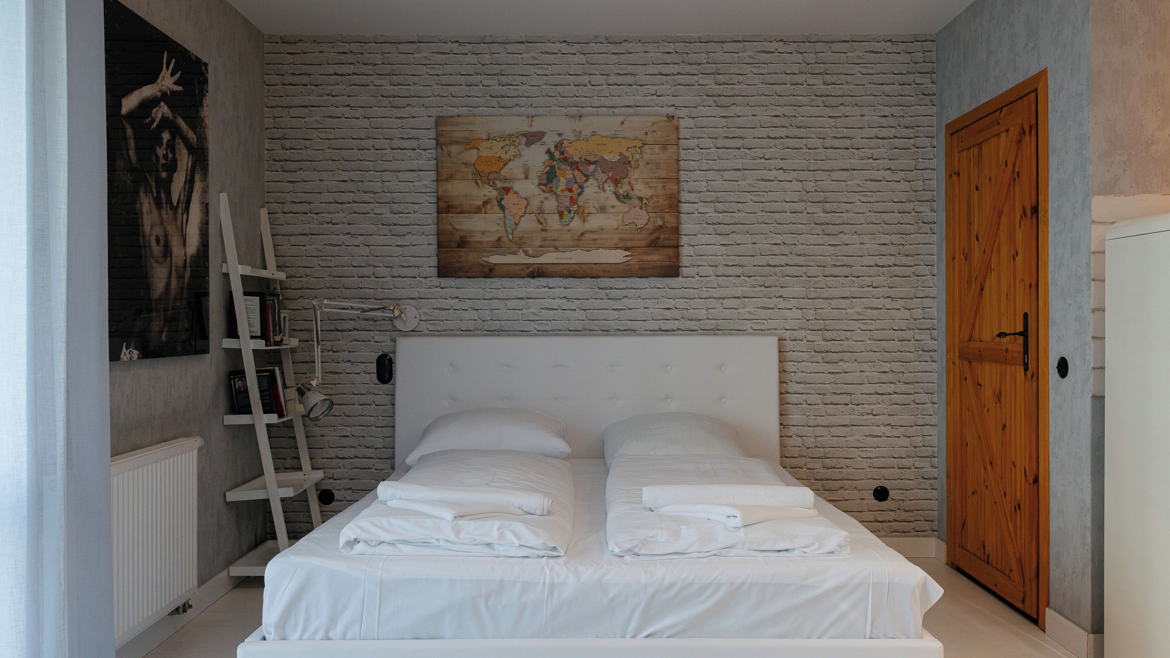 A bedroom with a white brick wall, a wooden door, a white bed with pillows, a wooden ladder, and a world map on the wall.